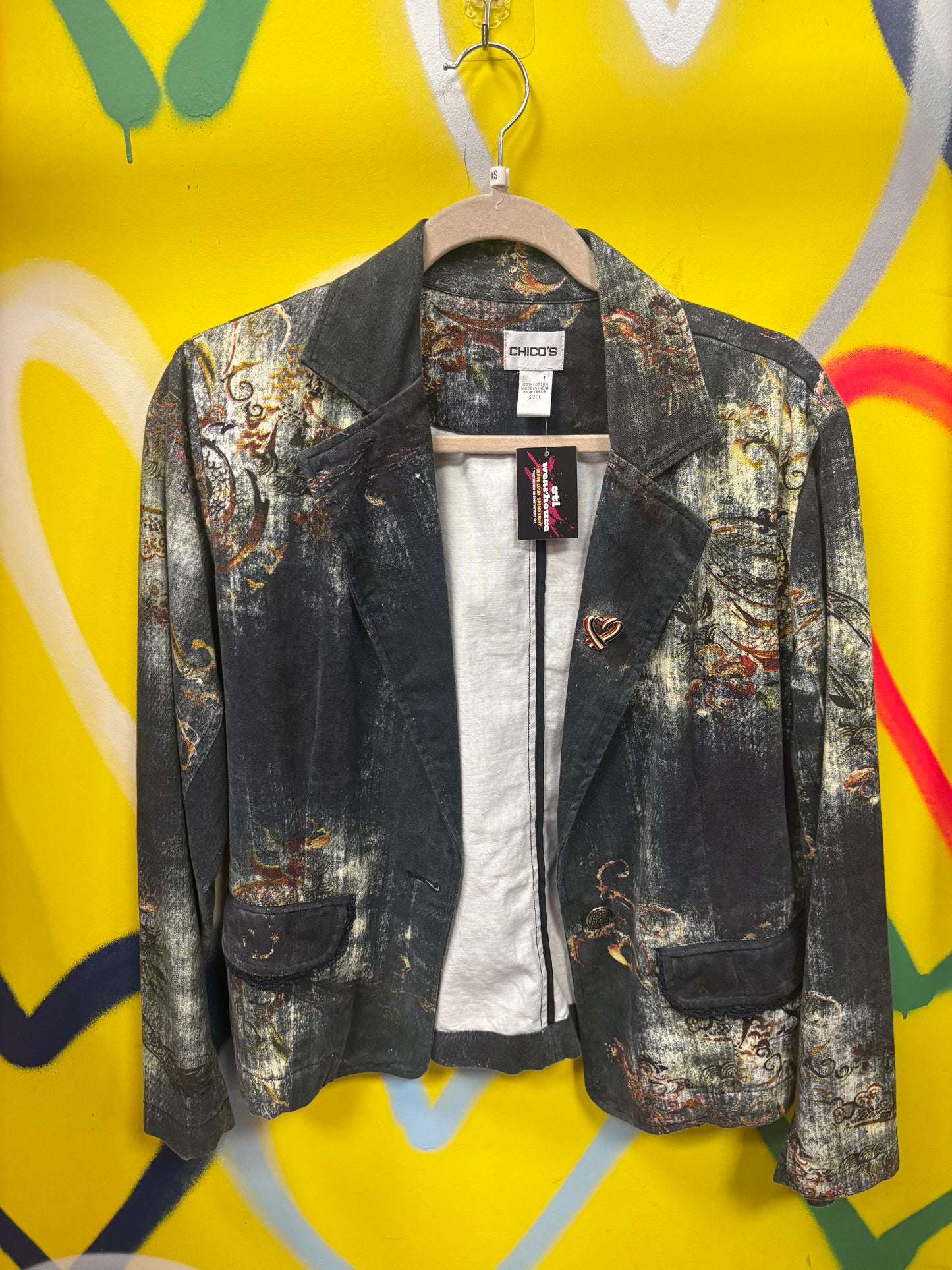 A dark gray and black distressed jacket with a colorful abstract print, hanging on a wooden hanger against a bright yellow graffiti-patterned background.