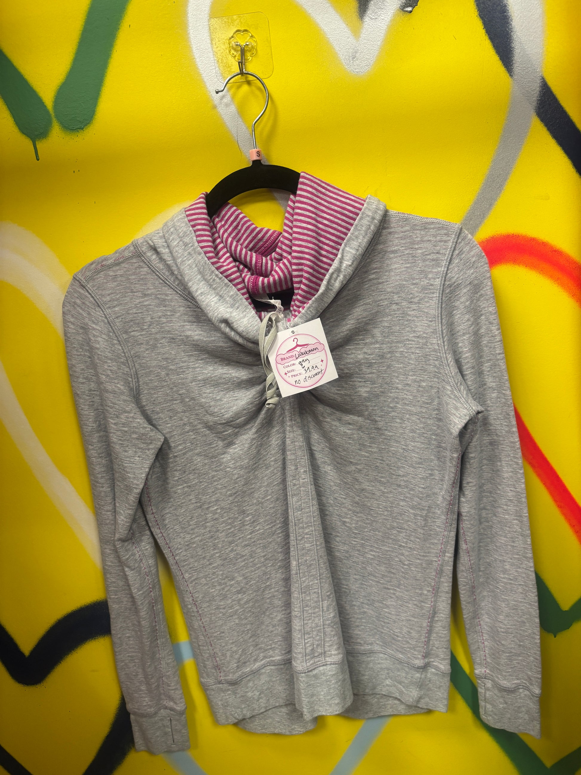 A gray long-sleeve hooded sweatshirt with a pink and white striped inner hood lining, hanging on a black hanger against a bright yellow graffiti wall. A white tag is attached to the collar.